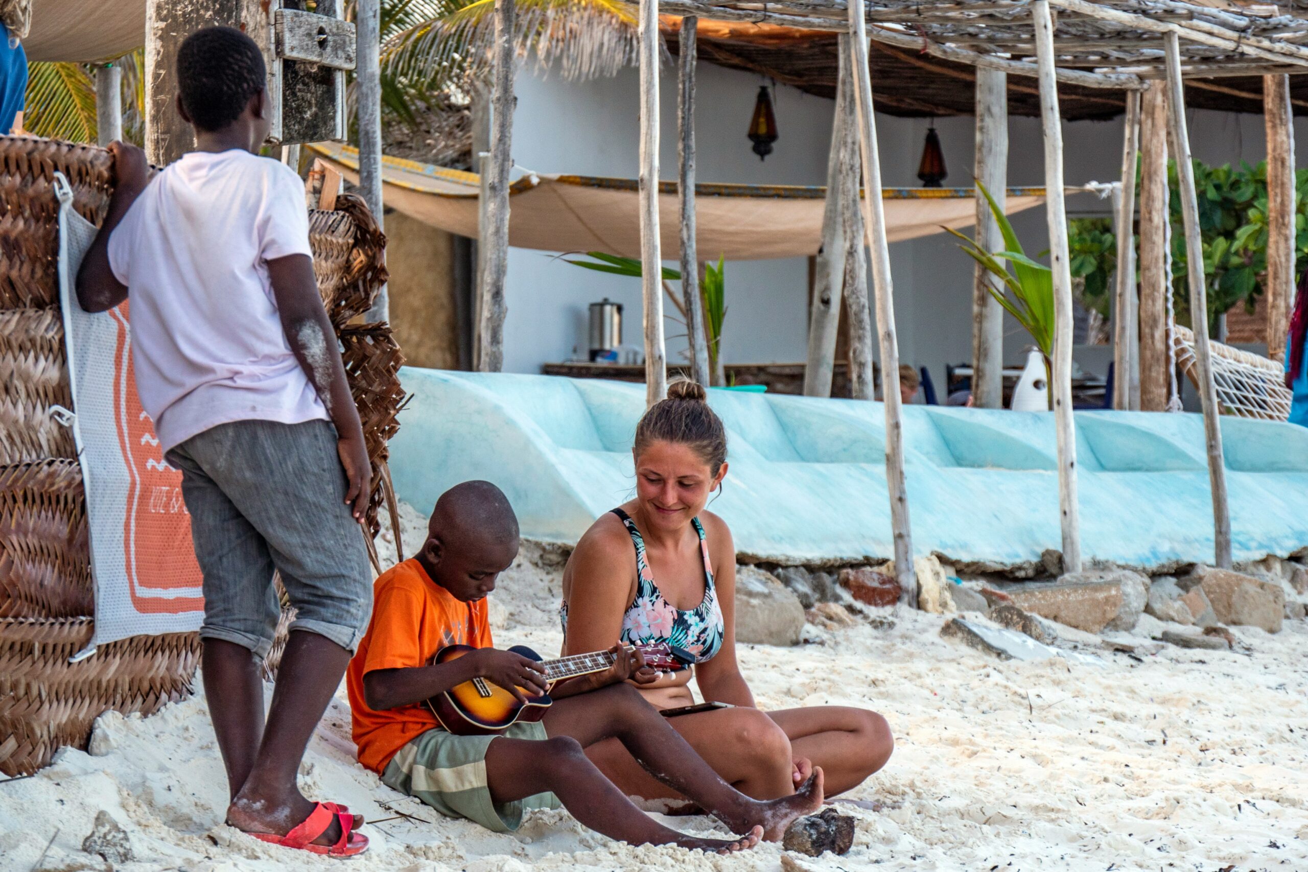 Traveler sitting on Paje Beach Zanzibar with local children, one playing a small guitar on the sand near beach huts